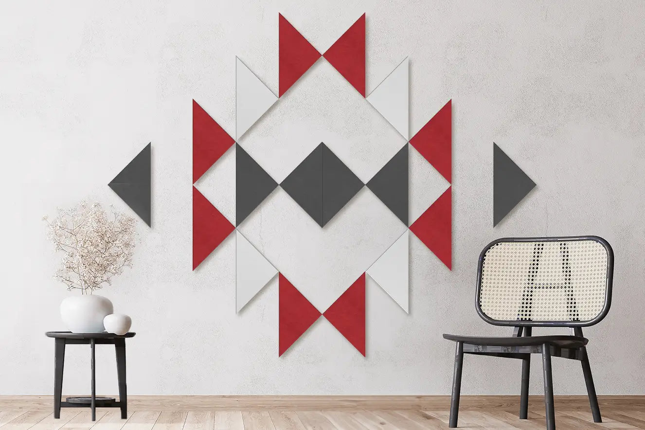 A geometric wall art set made of red, gray, and white triangular panels arranged in a diamond pattern on a light gray textured wall, with a black chair and a small side table in a styled room.