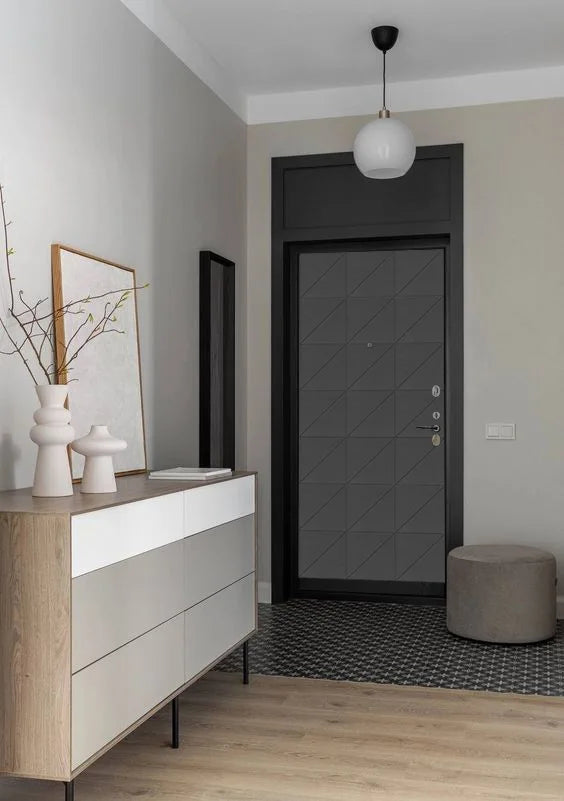 A modern entryway featuring a dark charcoal gray geometric-patterned front door, a light wood and white sideboard cabinet, a gray cylindrical ottoman, and a black-and-white patterned tile runner.