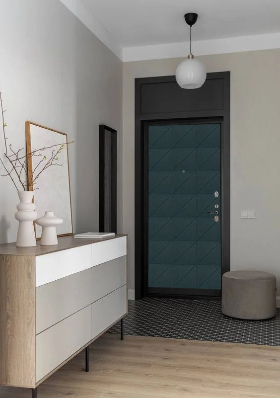 A modern entryway featuring a dark teal geometric-patterned front door with a black frame, a light wood and white dresser, a white pendant light, and a gray cylindrical ottoman on a black-and-white patterned rug.