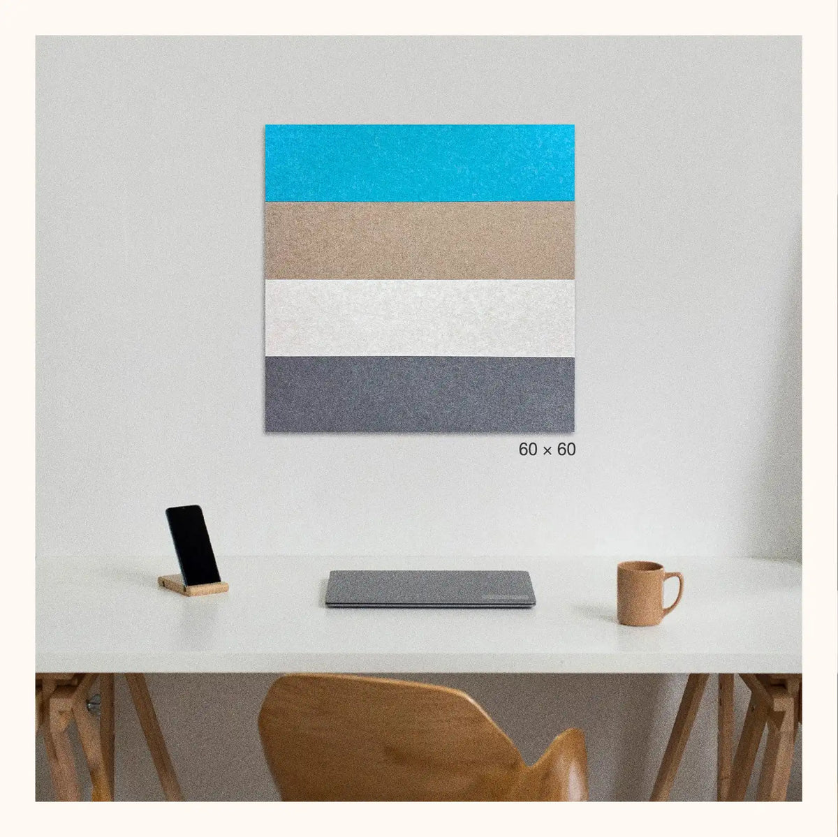 A square wall art panel with four horizontal color bands bright blue, tan, white, and dark grey is displayed above a white desk in a minimalist room setting. The print is shown on a light gray wall, with a smartphone, tablet, and mug on the desk below.