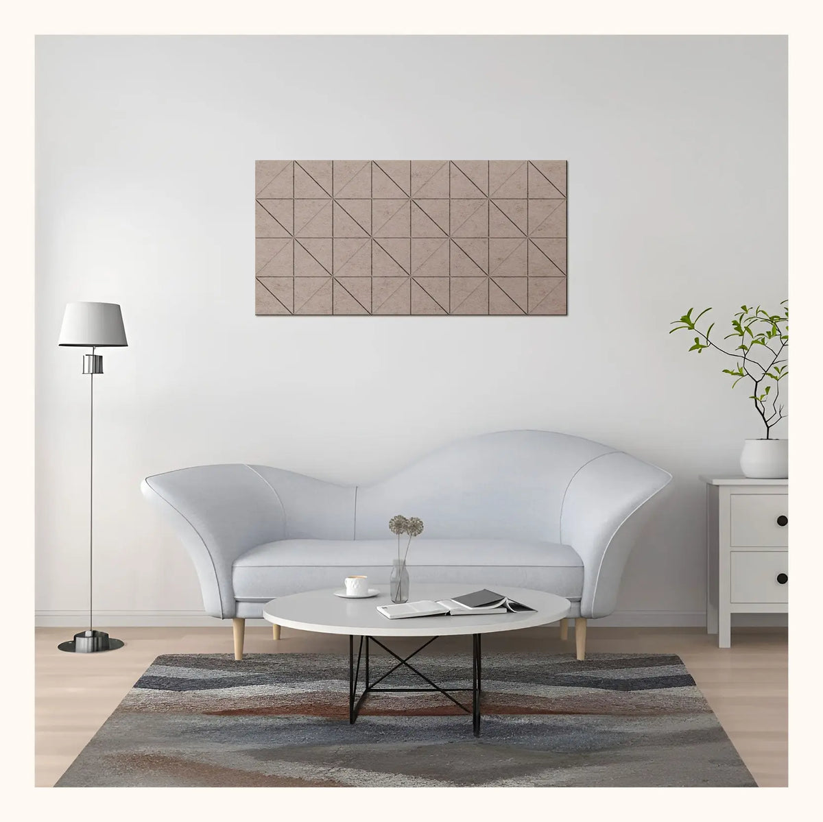 A modern living room scene featuring a rectangular wall art piece with a geometric triangular pattern in beige and brown tones, hung above a light gray curved sofa.