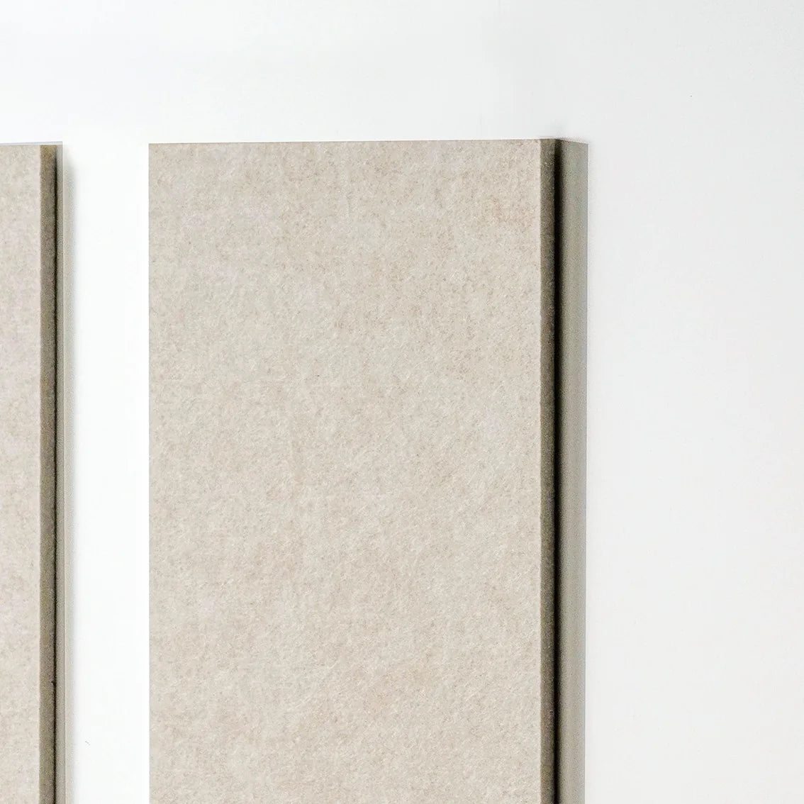 A close-up of a rectangular wall art panel with a textured light tan surface, shown against a plain white wall. Part of a second panel is visible at the left edge.