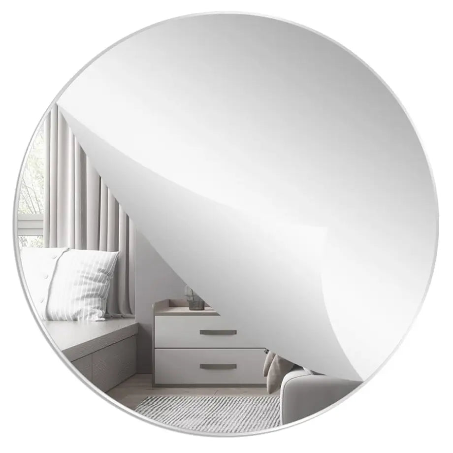 A round convex mirror with a silver frame, shown on a white background with a curled-back top edge revealing a glossy finish. The mirror reflects a modern gray bedroom interior with a window, bench, and drawers.