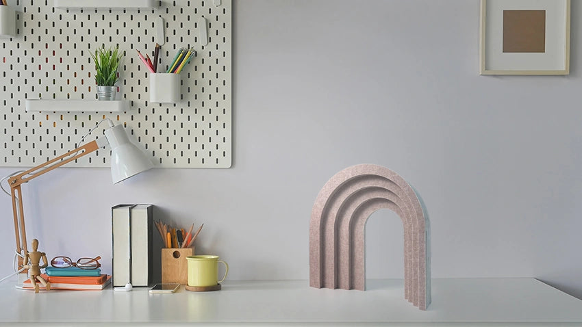 A beige felt arch bookend with concentric arch layers stands on a white desk in a styled home office setting next to a desk lamp, books, and a mug.