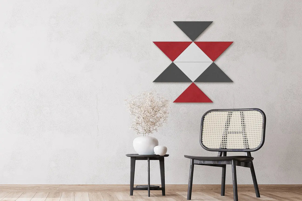 A geometric wall art piece made of red, dark gray, and white triangles mounted on a textured light gray wall above a black side table and a woven-back chair.