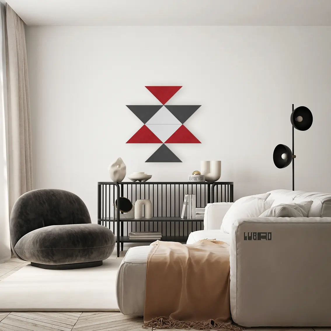 A modern living room interior featuring a geometric wall art piece made of red, gray, and white triangles mounted above a black wire console. The room includes a white sofa with a beige throw blanket, a dark gray velvet lounge chair, a black floor lamp, and light beige curtains.