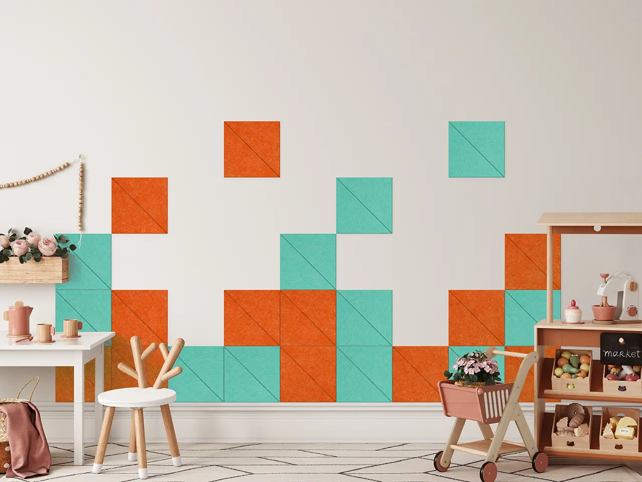 A styled children's room corner featuring a wall decorated with orange and teal geometric square tiles. The room includes a white table with a wooden stool, a wooden toy cart with flowers, and storage baskets on a light wood shelf.