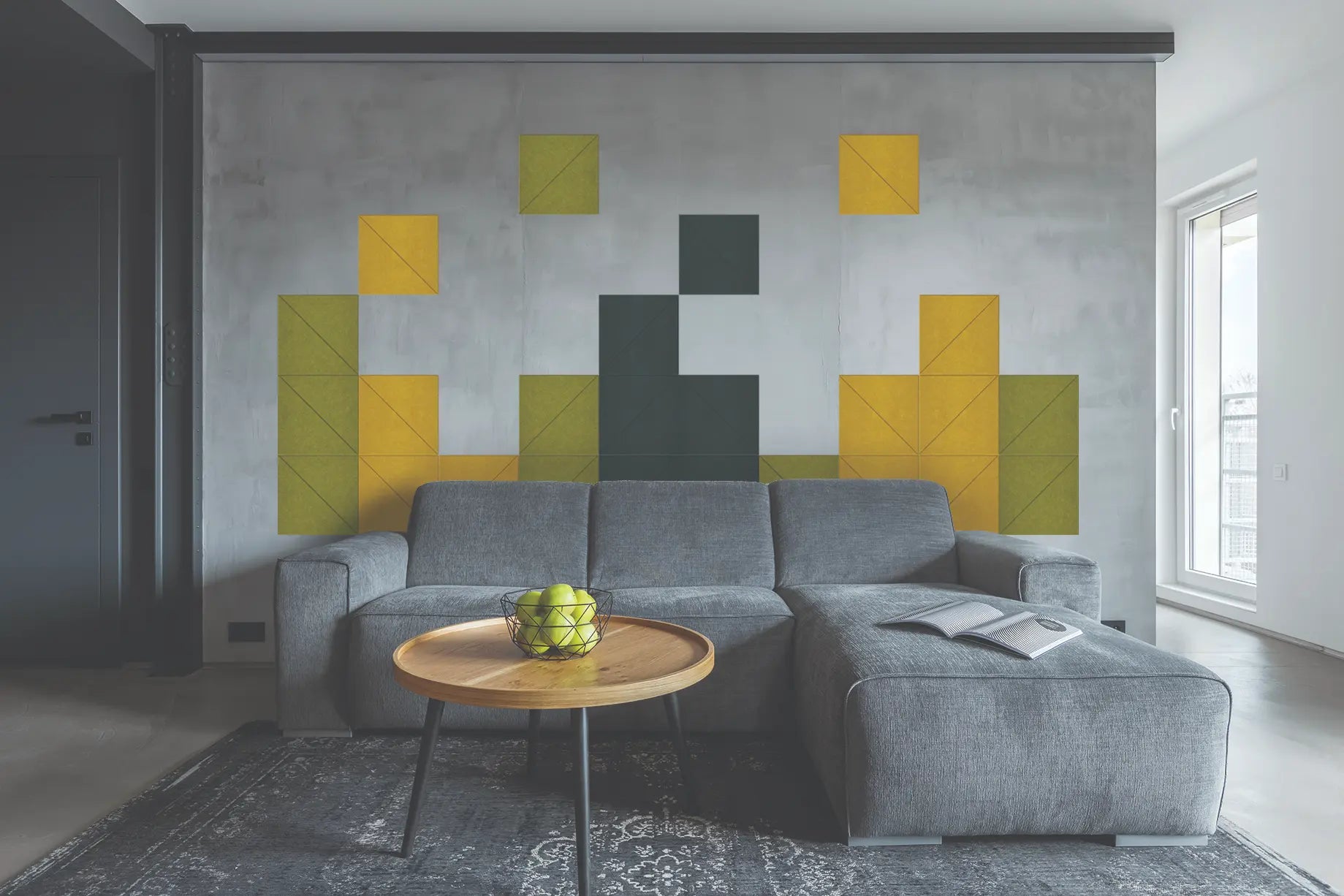 A modern living room featuring a large gray concrete accent wall with a geometric pixel-art style pattern in yellow, olive green, and dark gray. In front of the wall is a gray sectional sofa with a round wooden coffee table holding a bowl of green apples.