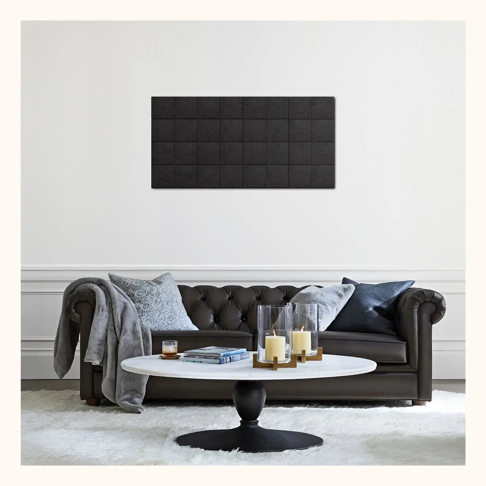 A modern living room scene featuring a black tufted leather sofa with gray and black pillows, a round white coffee table with candles, and a black rectangular wall panel mounted on a white wall.
