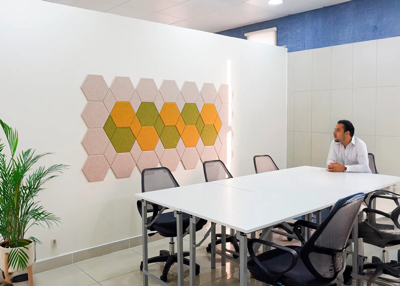 A modern meeting room with a white conference table, black mesh office chairs, and a man sitting at one end. On the white wall behind the table is a decorative arrangement of hexagonal acoustic panels in beige, green, and orange.