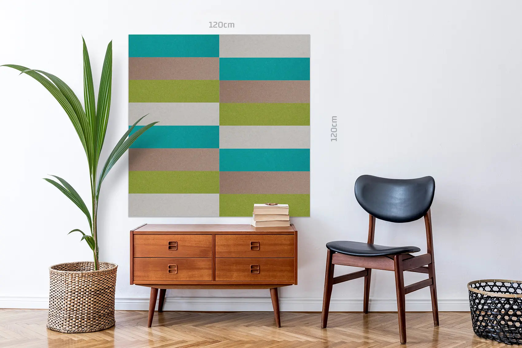 A large square wall art print with a geometric stripe pattern in teal, olive green, beige, and brown hangs on a white wall in a styled room. A mid-century wooden cabinet, black chair, potted plant, and basket are arranged below.