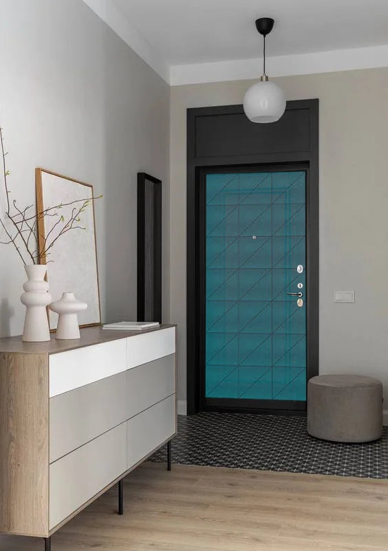 A modern interior entryway featuring a teal blue security door with a black geometric grid pattern, set in a dark frame. The door is styled in a contemporary room with a light wood floor, a gray cylindrical ottoman, a white and beige sideboard cabinet, and a white globe pendant light.