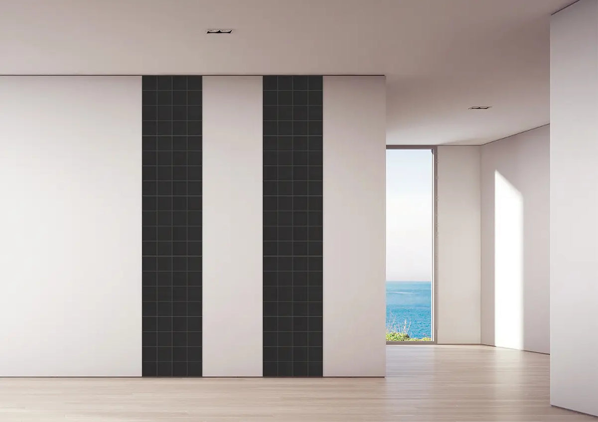 Modern interior wall paneling featuring two tall vertical black grid-patterned panels flanked by white panels, set in a minimalist room with light wood floors and a window looking out to the ocean.